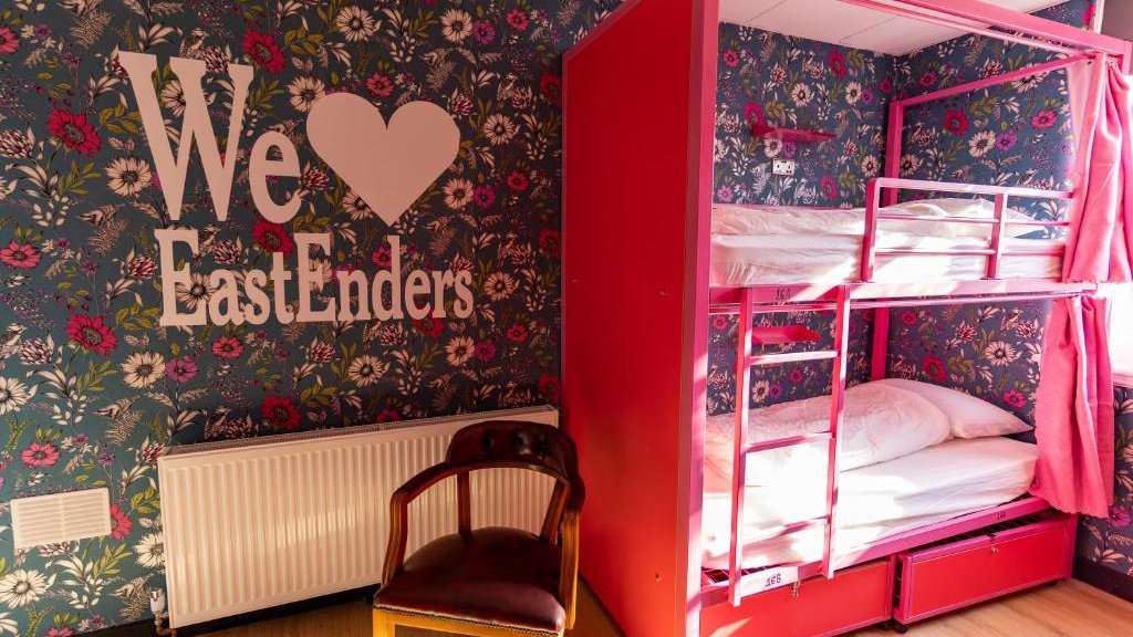 Two pink bunk beds in a room with floral wallpaper and "We <3 Eastenders" in white on the wall.