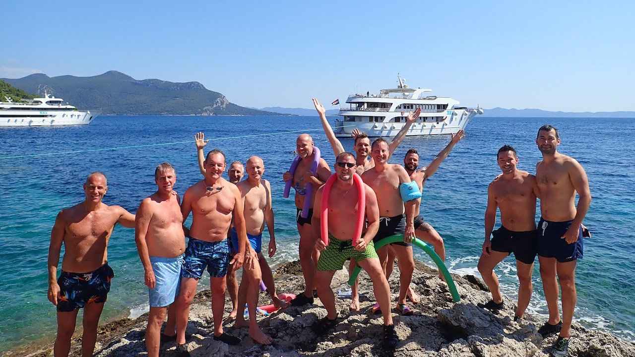 A group of men in shorts posing on a rocky outcrop with a luxury yacht in a bay behind them.