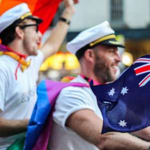 Two men in sailor outfits waving Australian and rainbow flags at a parade.