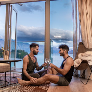 Stefan and Seby sitting on the floor of a hotel room sharing glasses of champagne with a view of a cloudy bay out the open door behind them.