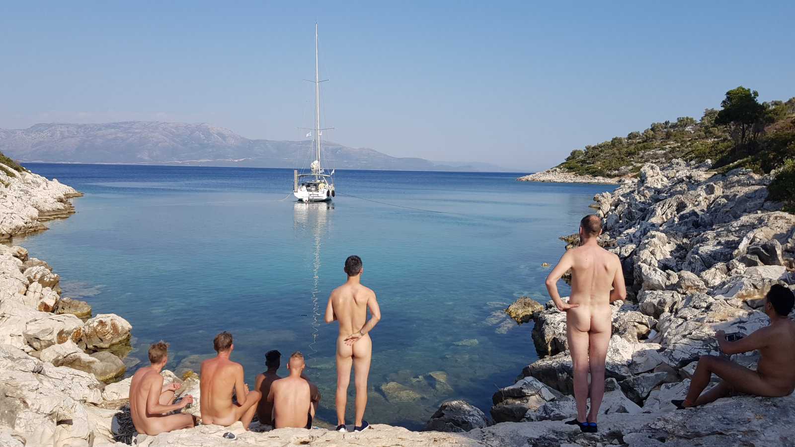 A group of nude men seen from behind as they look out over a yacht moored in a rocky bay.