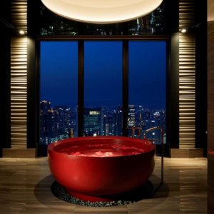 A big round red bathtub inside a room with massive windows overlooking a city at night.
