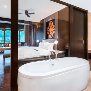 A big white bathtub next to a window showing a hotel bed in a wood paneled room.