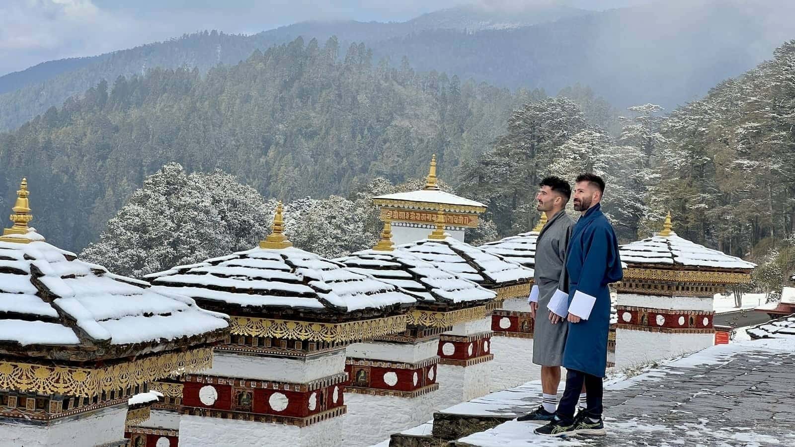Gay couple wearing a gho at the Dochula Pass with a blanket of snow covering the stupas.