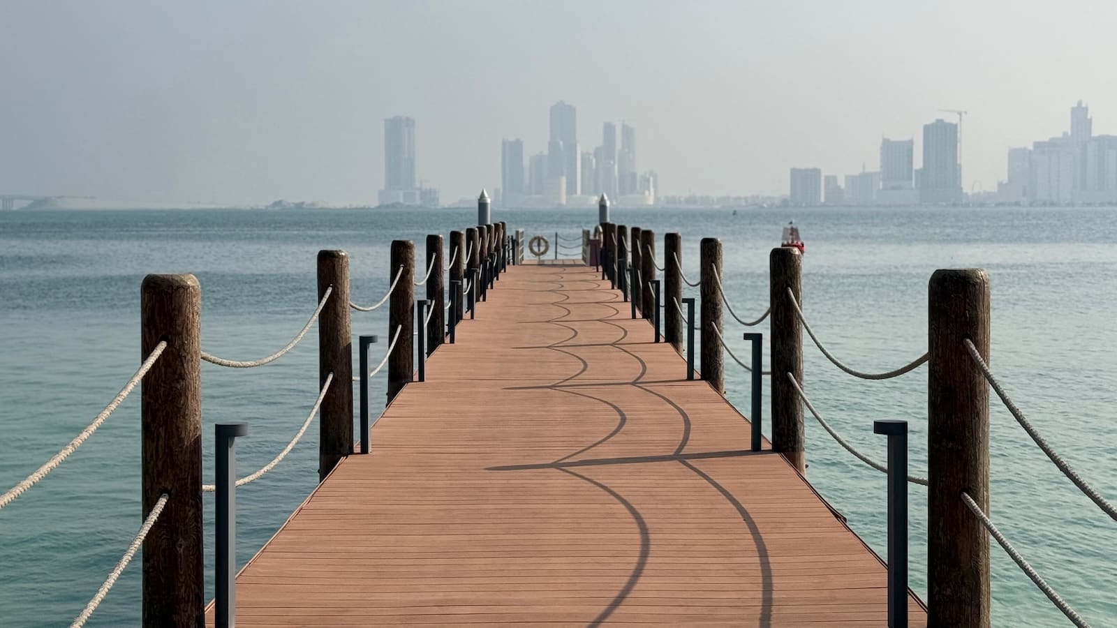 A jetty pointing towards a hazy city skyline across some water on a sunny day.