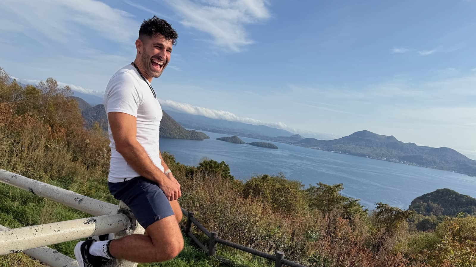 Stefan sitting on a fence and laughing with a spectacular bay and greenery behind him.