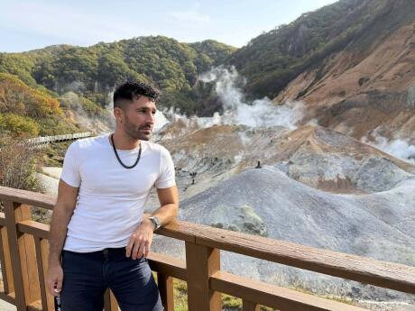 Stefan leaning against a fence and looking away from the camera with hot springs behind him.