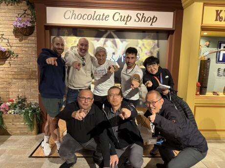 Stefan and a group of men all pointing at the camera as they pose in front of a shop sign that says "Chocolate Cup Shop."