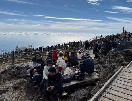 A crowd of people sitting on top of a mountain peak looking down over clouds.