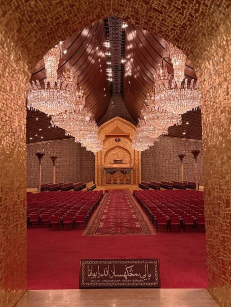 An opulent hall with sparkling chandeliers and red seats set before a golden altar.