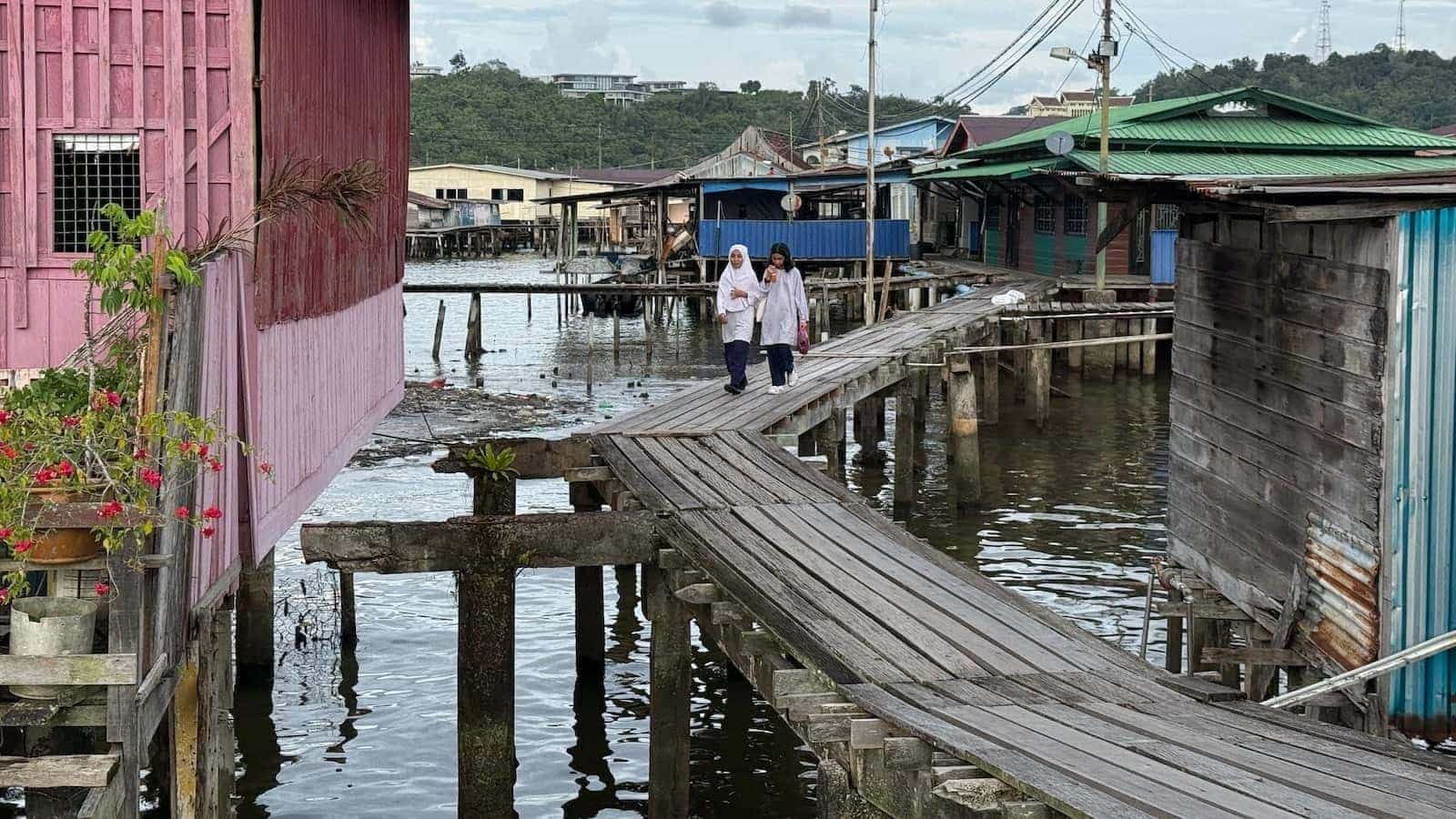 Two young girls walking across raised platforms above water with houses on stilts around them.