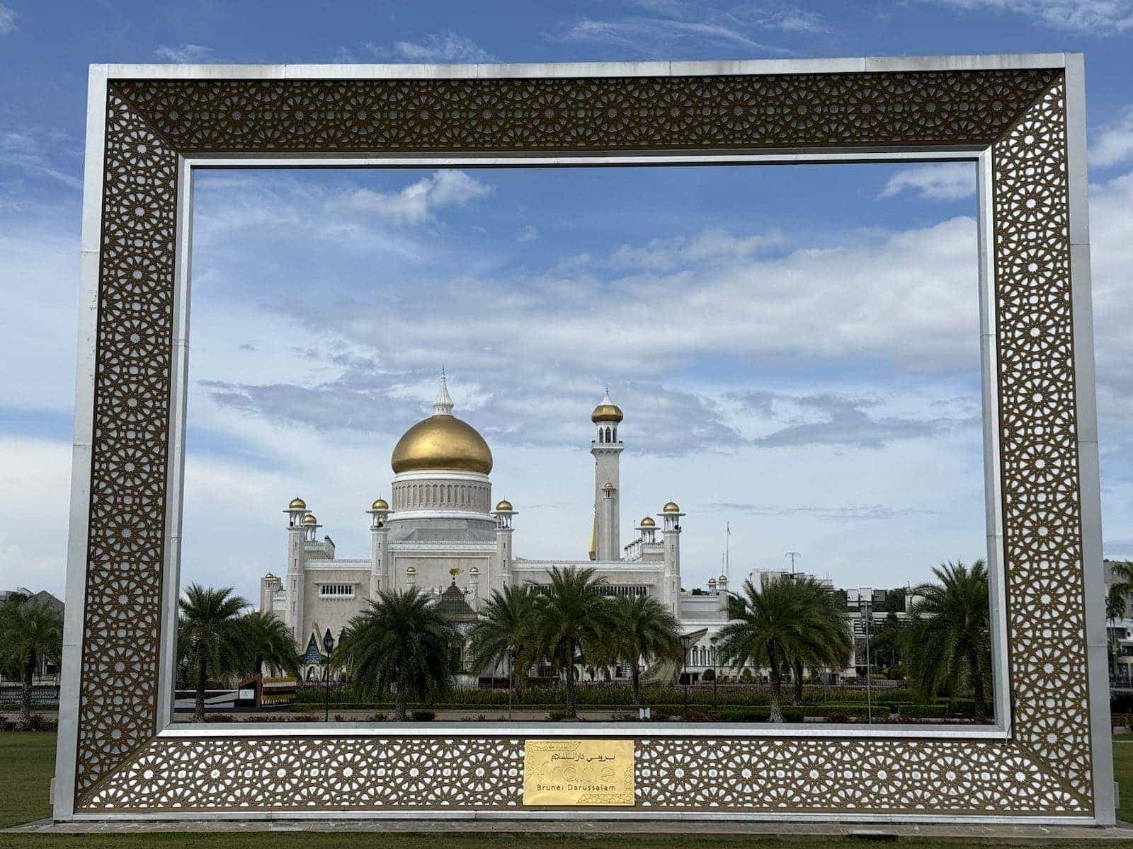 A white mosque topped with golden domes photographed through a frame from a distance away.