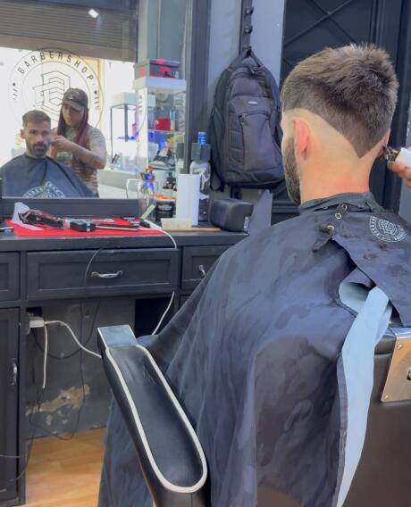 Seby sitting in a barber's chair getting a haircut as seen from behind, and in the reflection of the mirror you can see his face.