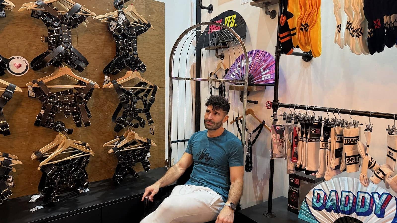 Stefan sitting in a chair in a gay shop, surrounded by harnesses and fans with cheeky messages on them.