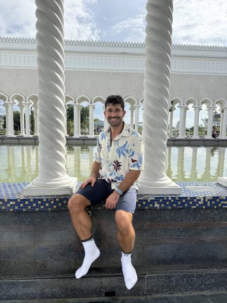 Stefan sitting on a wall in an outdoor courtyard of a mosque with columns and water behind him.