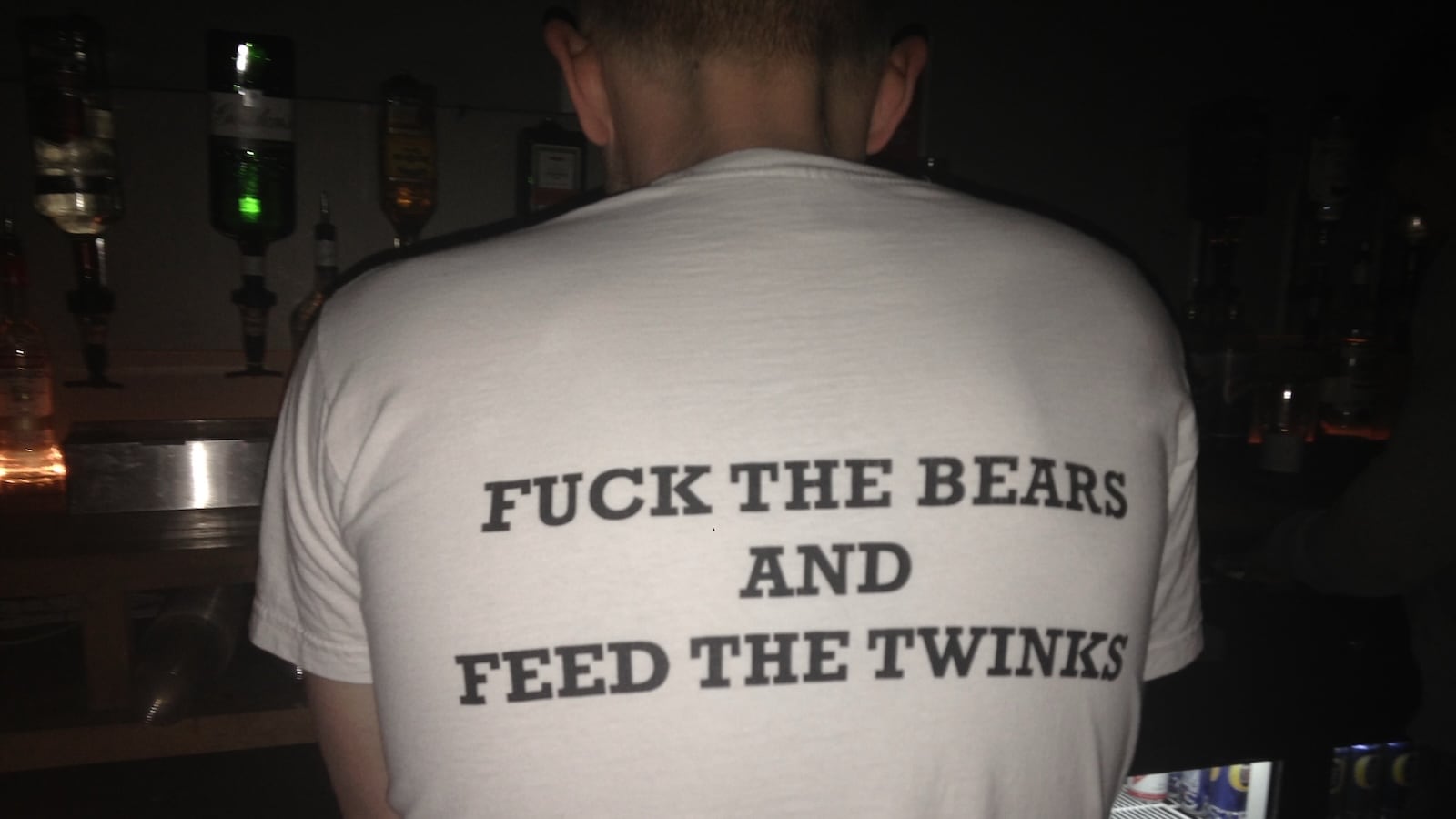 A man standing with his back to the camera wearing a t-shirt that says, "fuck the bears and feed the twinks."