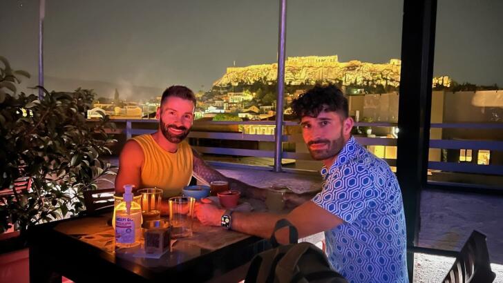 Gay couple Stefan and Seby having a romantic cocktail at a rooftop bar in Athens with view of the Acropolis.