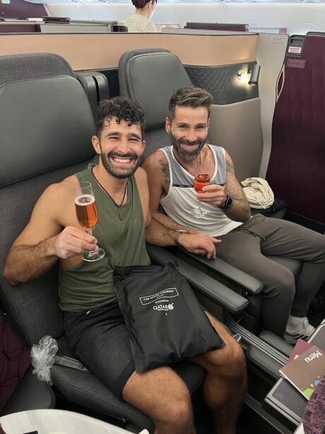 Stefan and Seby posing with glasses of champagne in airline seats on a Qatar Airlines plane.