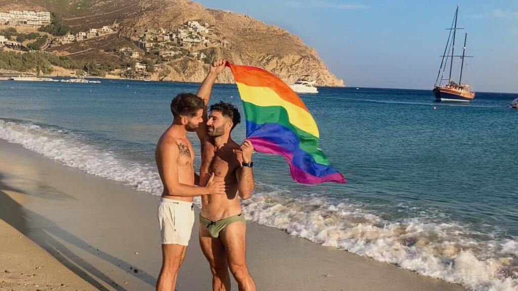 Stefan and Seby holding a rainbow flag on a beach with a yacht in the background.