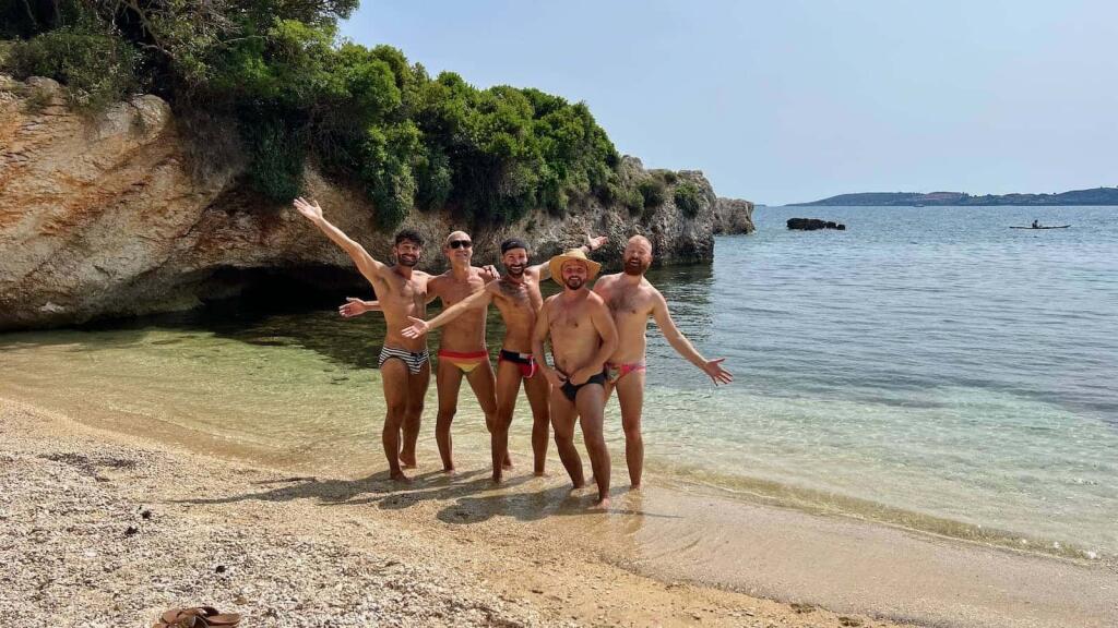 Five men in speedos posing on a beach in the sun.