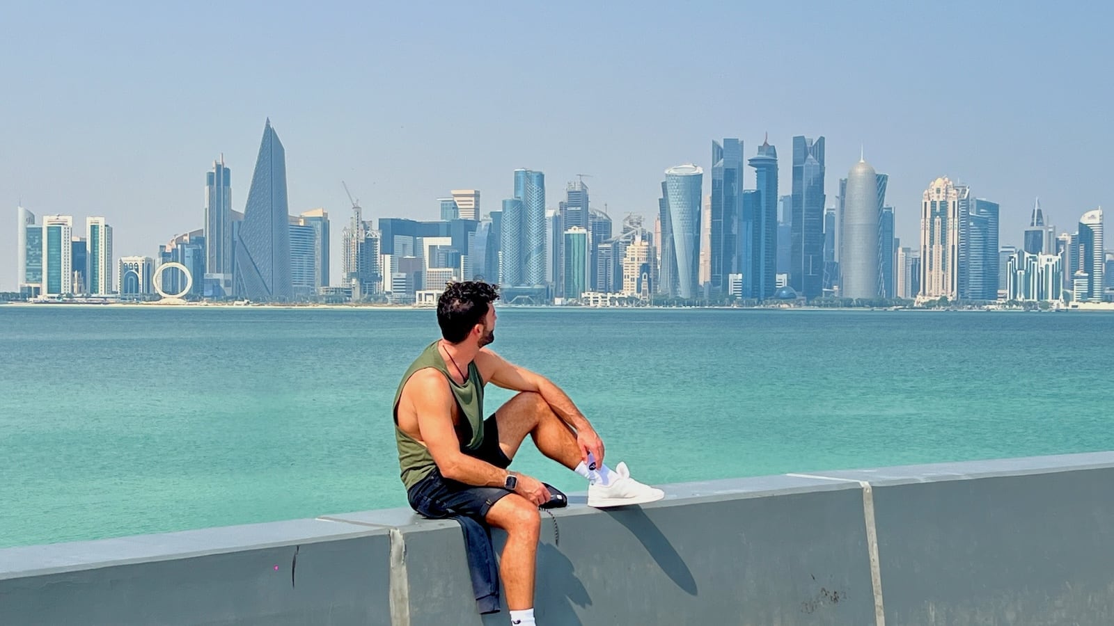 Stefan sitting on a wall with brilliantly blue water and skyscrapers in the background.