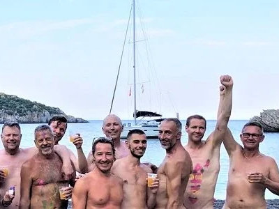 A group of nude men posing on a beach with a yacht in the background.