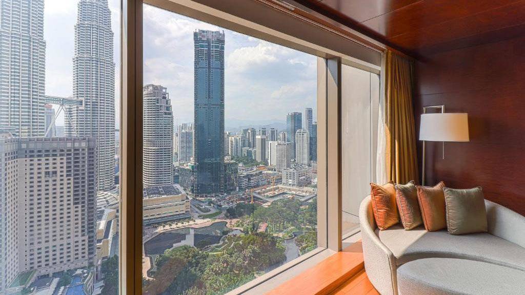 A rounded couch in a hotel room next to huge windows with a view of gardens and skyscrapers outside.