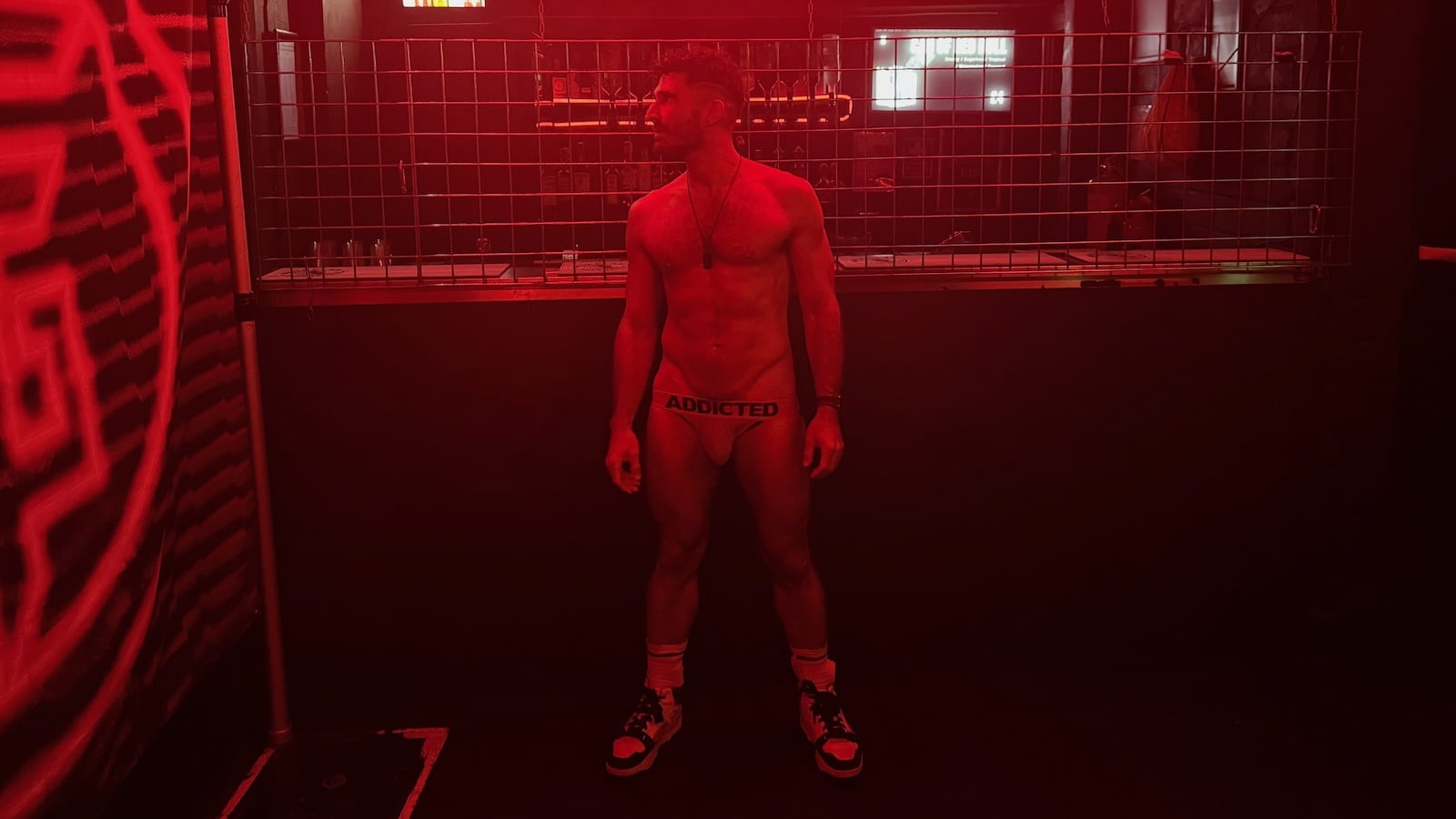 Stefan standing in a club lit by red light in nothing but briefs and sneakers.