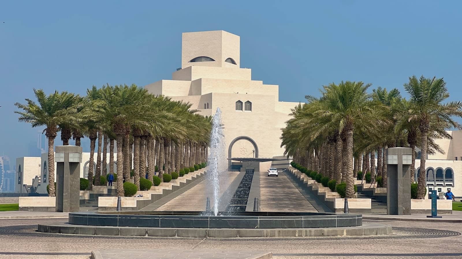 A white building made of squares at different angles with palm trees and a fountain in front.