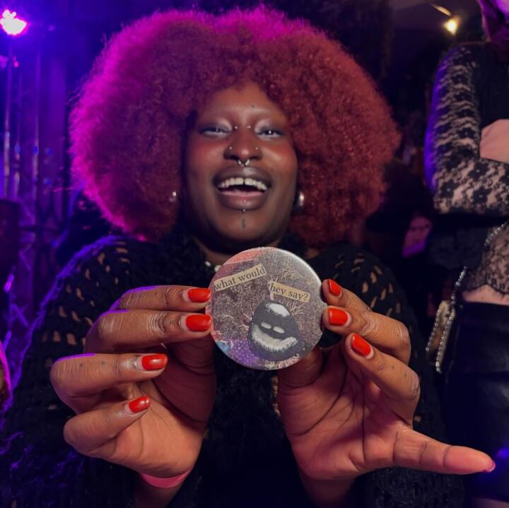 A pierced black woman with an afro and red nails holding up a badge that says, "what would they say?"