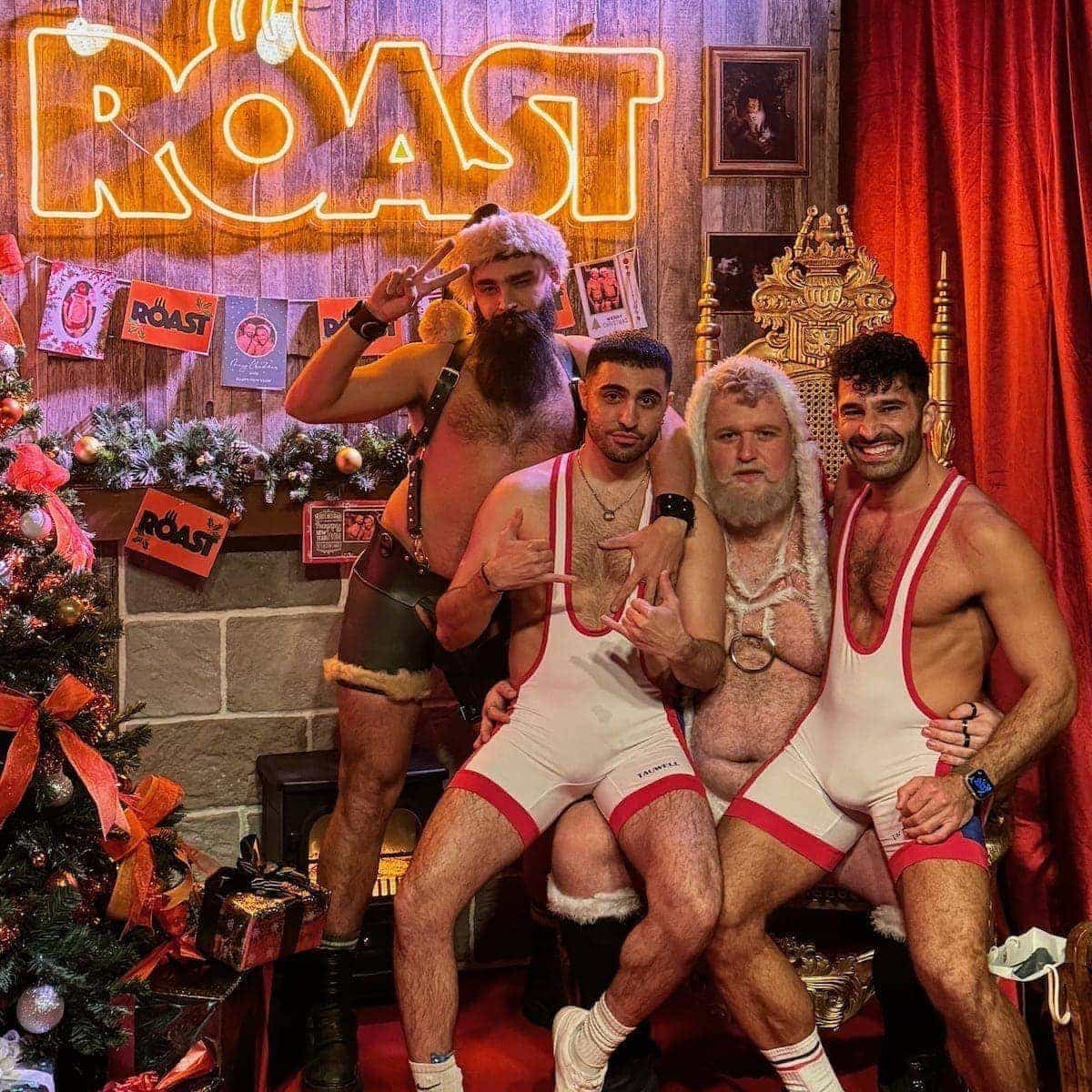 Stefan and a friend sitting on a gay Santa's knee in matching outfits with another bearded man and a sign saying ROAST behind them.