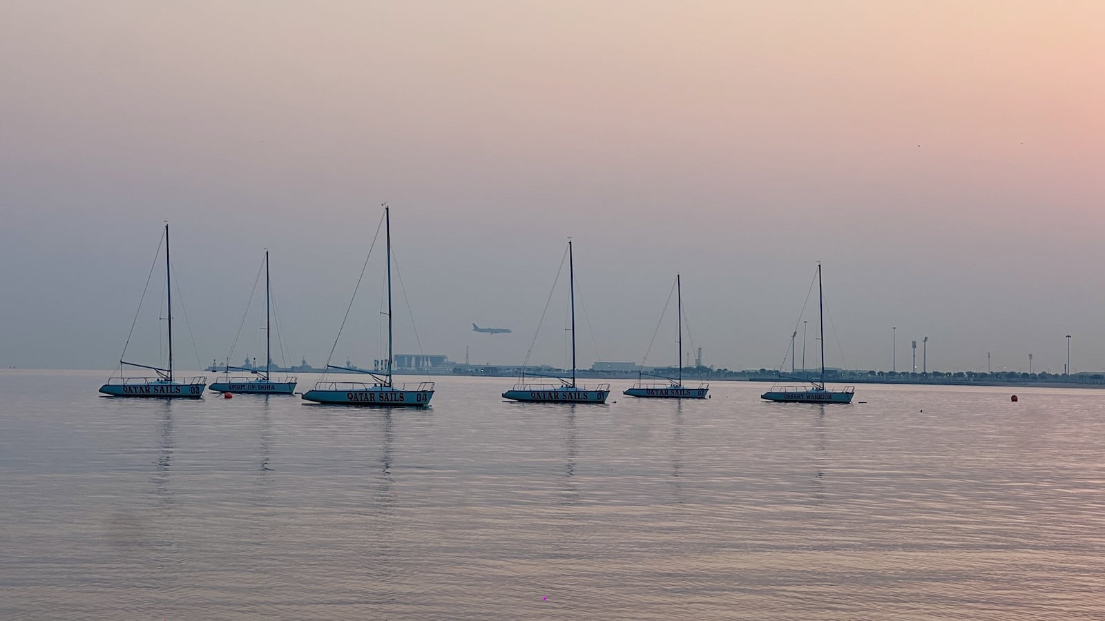 Six yachts silhouetted on the water with a plane landing in the far distance on land in the background.