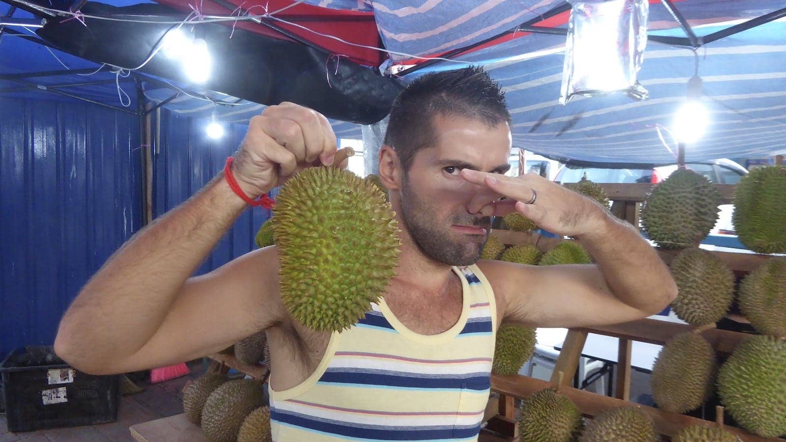 Seby in a night market holding a durian fruit in one hand and pinching his nose closed with the other!