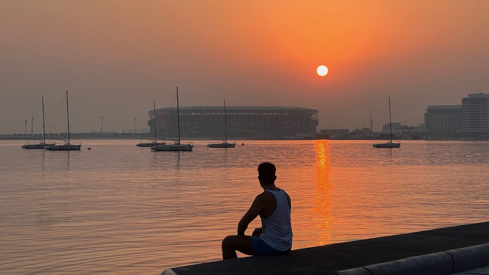 Stefan silhouetted sitting next to water with yachts and an orange sun in the background.