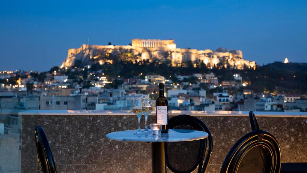 An Athens hotel balcony at night with chairs, a table and wine, plus a stunning view of the Acropolis lit up at night in the background.