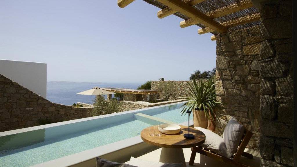 View from a stone hotel balcony looking over a small infinity pool to the ocean on a sunny day.